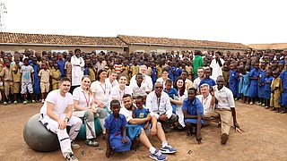 Deutsche Handwerker im Hof einer afrikanischen Schule sind umringt von afrikanischen Schulkindern in blauen Schuluniformen.