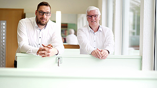 Zwei Männer, angelehnt auf einer halbhohen grünen Wand.