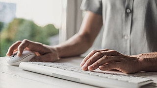 Eine Person tippt mit der linken Hand auf einer Tastatur und hat in der linken Hand die Computermaus