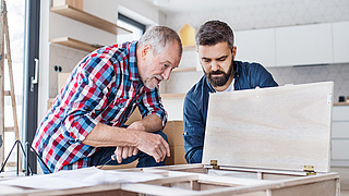 Ein älterer und ein jüngerer Tischler bauen Küchenschränke ein.