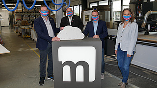 Drei Herren und eine Dame mit blauen Masken in einer Tischlerei-Werkstatt.